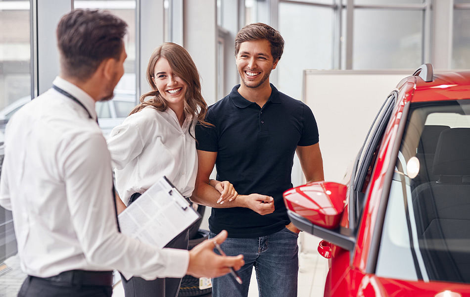 A man and woman engage in conversation with a car salesman at a dealership, discussing vehicle options and pricing.