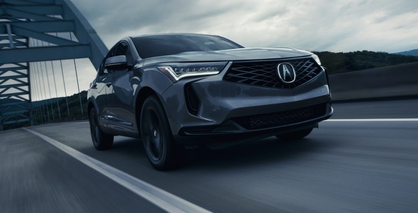 A sleek gray 2025 Acura RDX drives on a highway with a modern bridge in the background, under a cloudy sky.