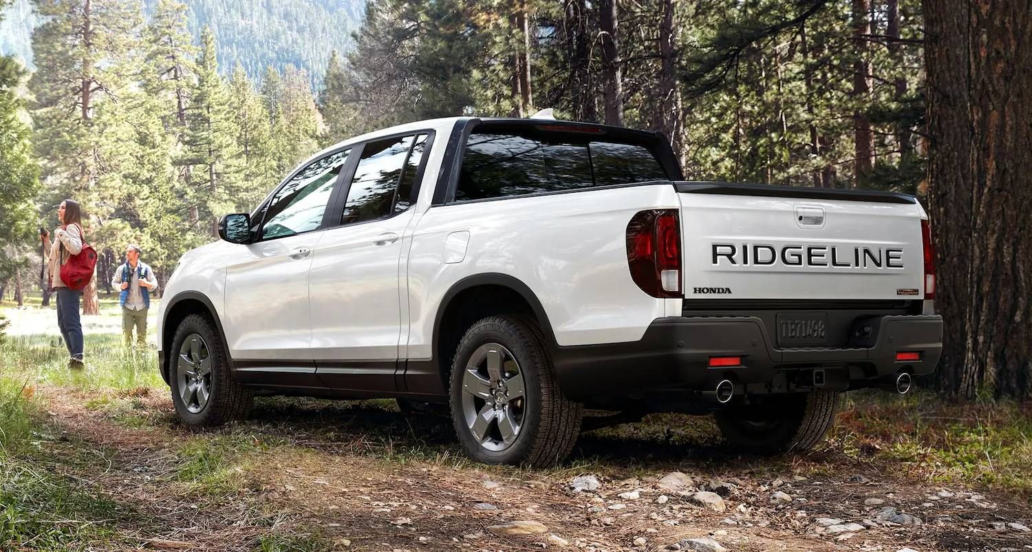 La Honda Ridgeline TrailSport 2025 en Platinum White Pearl estacionada junto a dos excursionistas en las montañas entre árboles altos