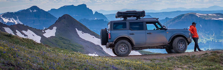 A person in an orange jacket stands beside their Ford Bronco on a mountain slope, gear loaded on the roof rack and ready for an adventure.