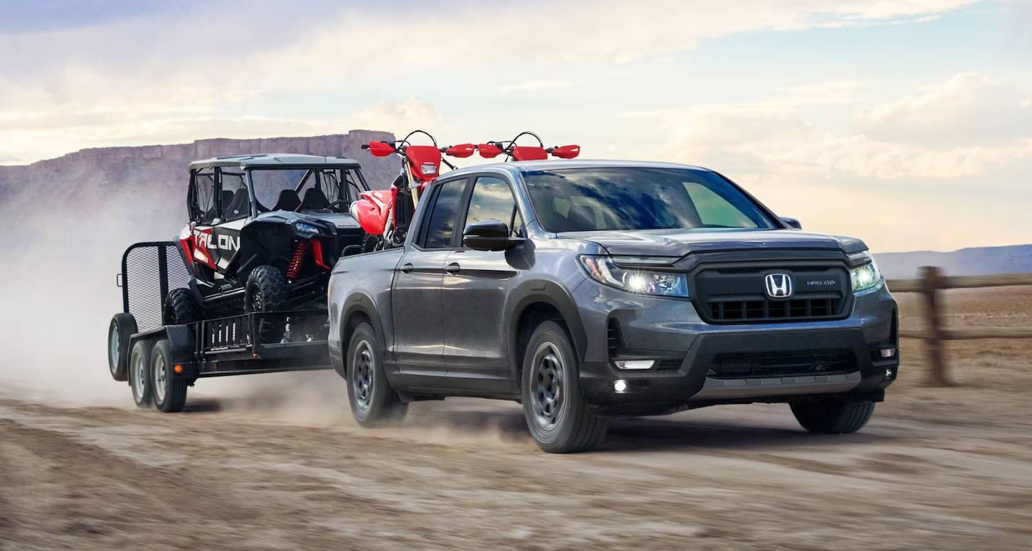 The 2025 Honda Ridgeline TrailSport in Modern Steel Metallic drives through a dirt field towing a trailer