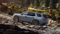 Un Honda Pilot TrailSport 2023 en color Sonic Gray Pearl con barras transversales accesorias y enganche para kayak circulando por un arroyo forestal.