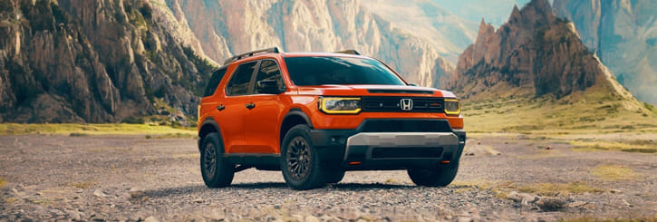 Un SUV de color naranja brillante con detalles en negro, estacionado en un terreno rocoso, con unas imponentes montañas escarpadas de fondo bajo un cielo despejado; transmite aventura y robustez.