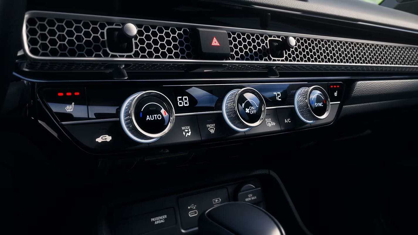 Close-up of a 2026 Honda Civic Hatchback Hybrid climate control panel featuring digital displays, knobs, and buttons. The modern design has a sleek, metallic finish and a sophisticated look.