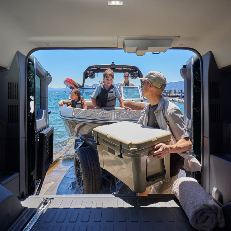  A view from inside the rear cargo area of an Ineos Grenadier looking out toward a sunny lake. A man is loading a large tan cooler into the trunk while a family waits on a boat docked at a ramp in the background.