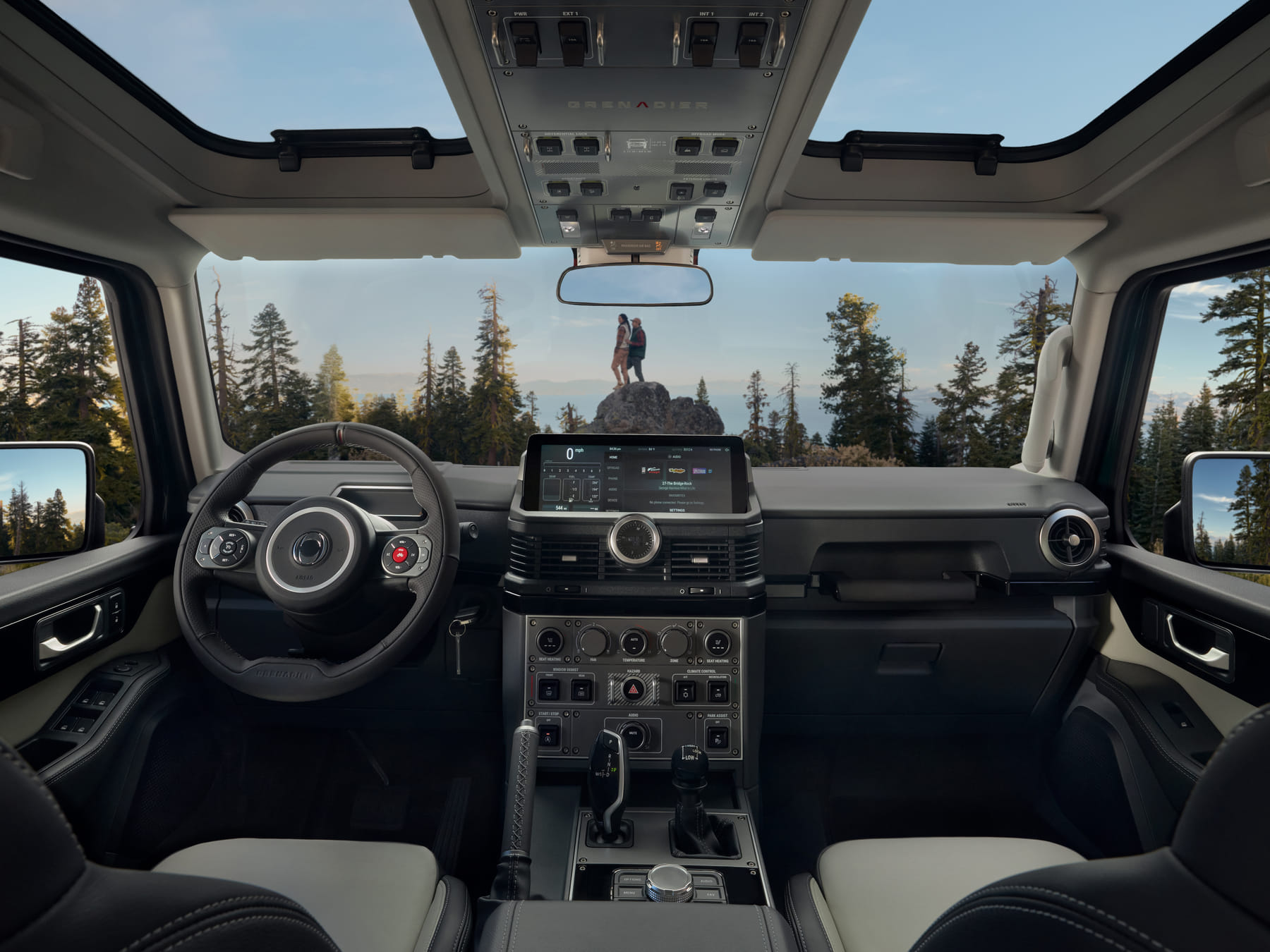 View from inside a rugged vehicle; dashboard in foreground with a forest visible through the windshield. Two people stand on a rock under a clear sky.