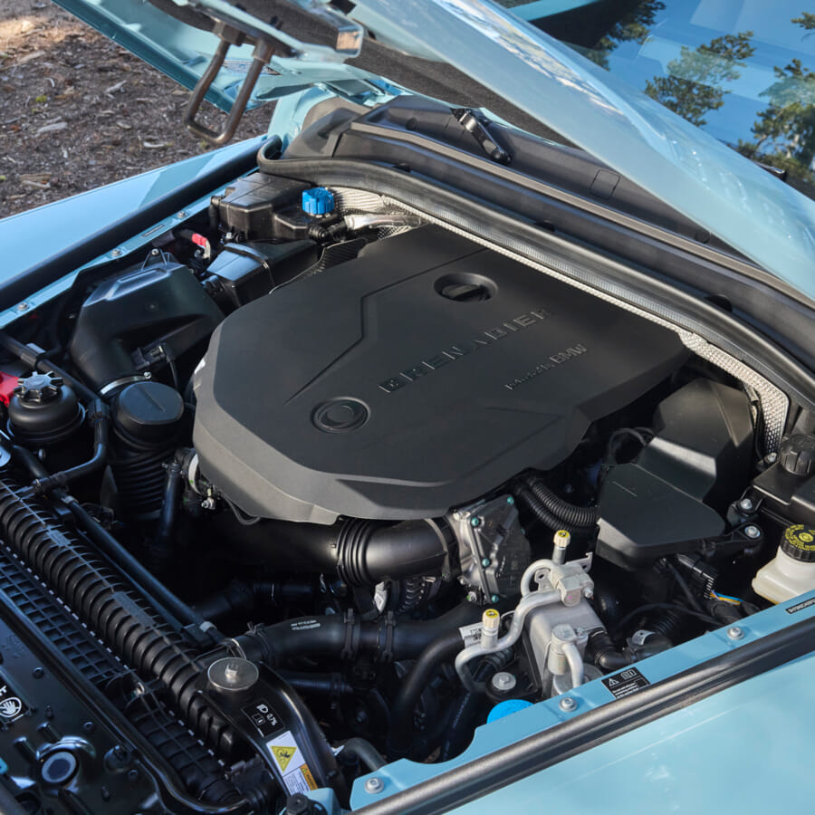 Car engine close-up under an open hood. The engine cover is sleek and black, with visible components surrounding it. Sunlight highlights the metallic parts.