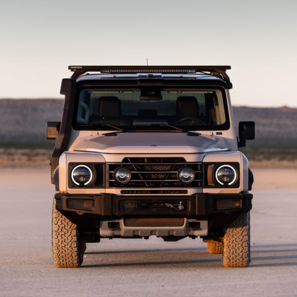 A rugged, silver SUV with round headlights and a roof rack stands on a flat, sandy landscape under a clear sky, conveying an adventurous feel.