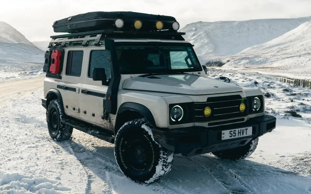 A modified white Ineos Grenadier Station Wagon parked on a snowy mountain road. The vehicle is equipped for off-roading with a rooftop tent, a roof rack with auxiliary lights, a snorkel, and a red side-mounted jerry can.