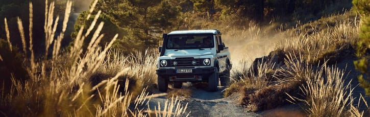 Ineos Grenadier Quartermaster drives through a dusty forest path surrounded by tall grass. The warm sunlight casts a golden glow, creating an adventurous tone.