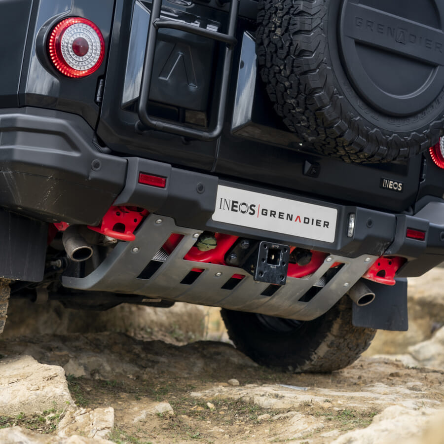Close-up of the rear of an INEOS Grenadier off-road vehicle on rocky terrain, showing robust tires, a spare tire, and protective underbody plating.