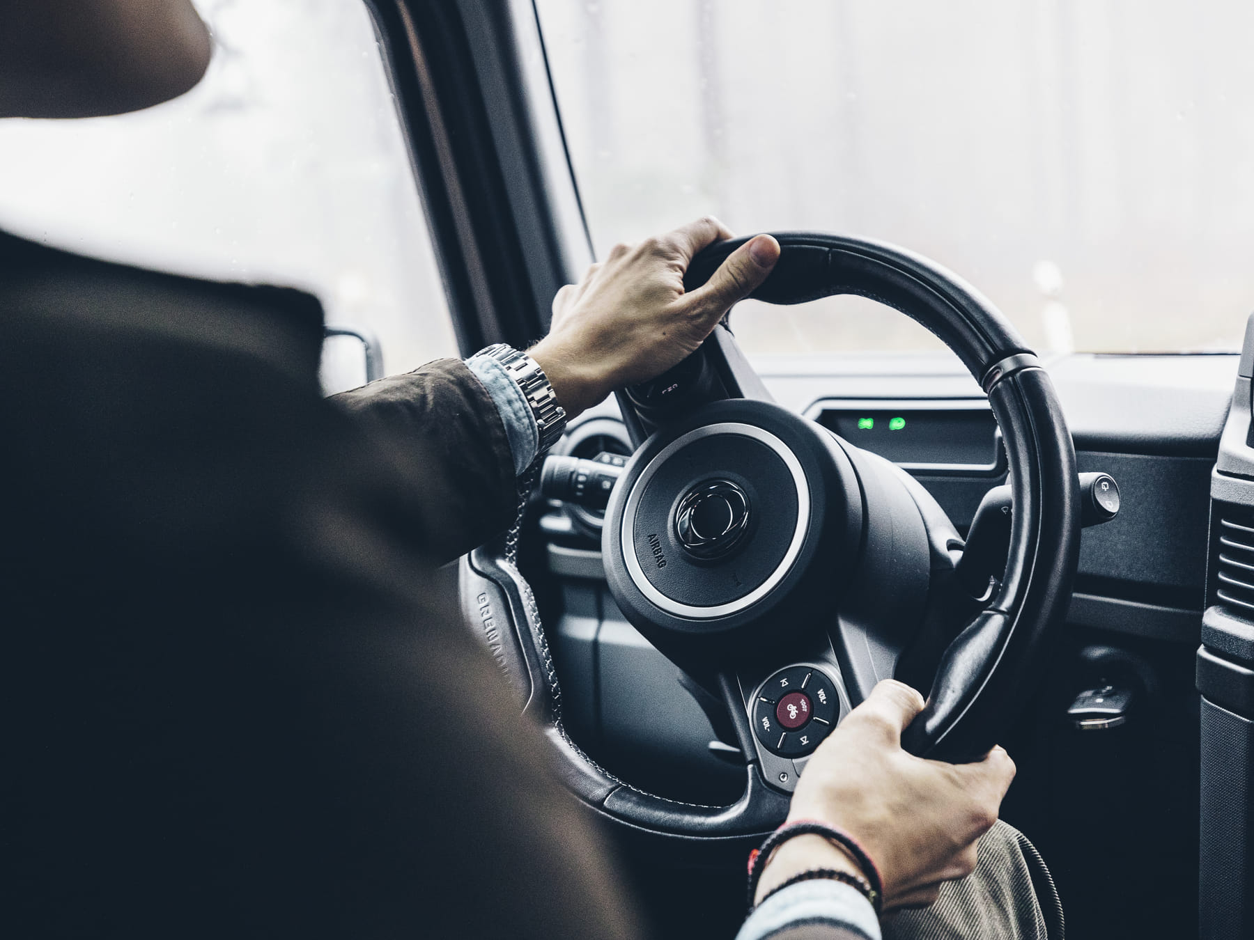 Close-up of a person's hands gripping a car steering wheel. The person is wearing a watch and bracelet, suggesting focus and control in driving.