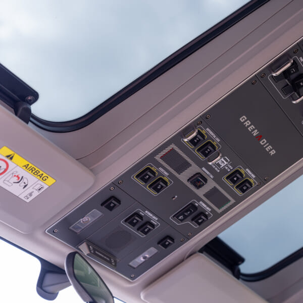 Close-up of the overhead control panel inside a vehicle, displaying various buttons and switches, labeled 'Grenadier.' The sunroof shows a cloudy sky.