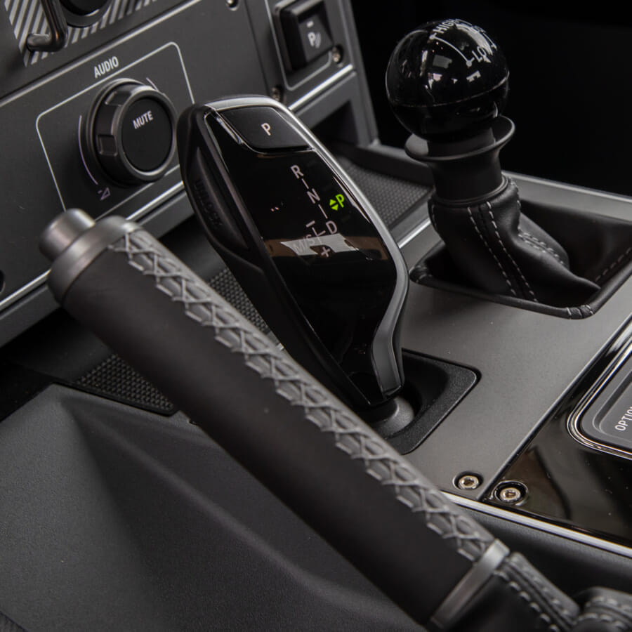 Close-up view of a car's gear shift area, featuring a modern gear selector, handbrake with stitched leather handle, and audio panel. Sleek and technical.