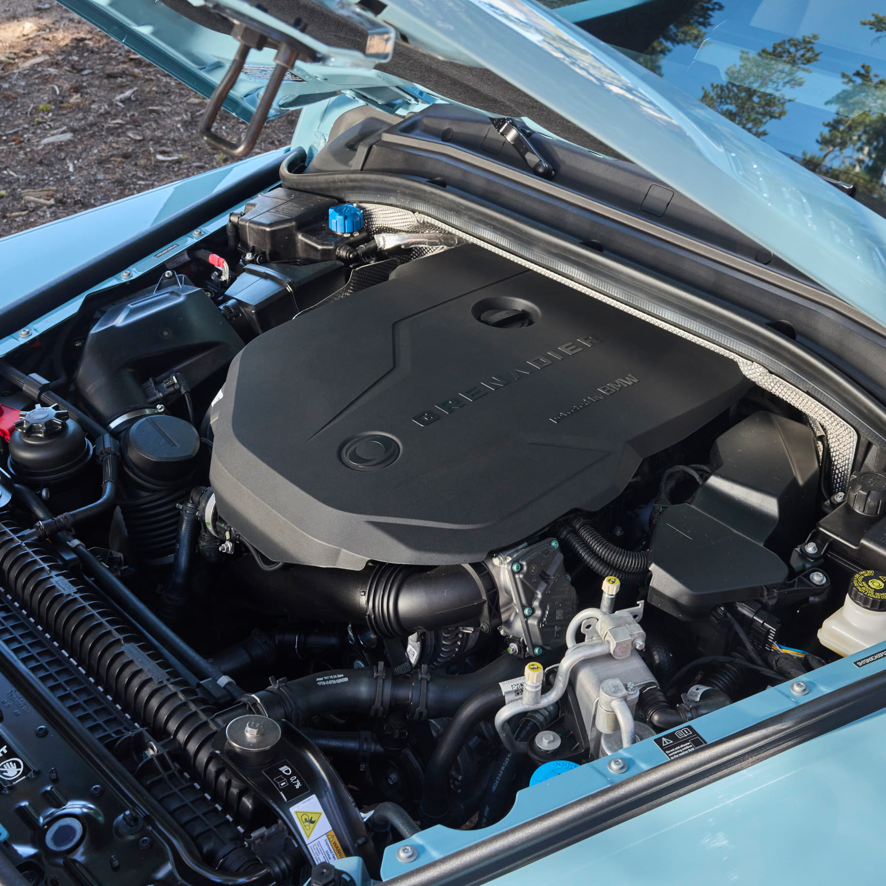 Car engine close-up under an open hood. The engine cover is sleek and black, with visible components surrounding it. Sunlight highlights the metallic parts.