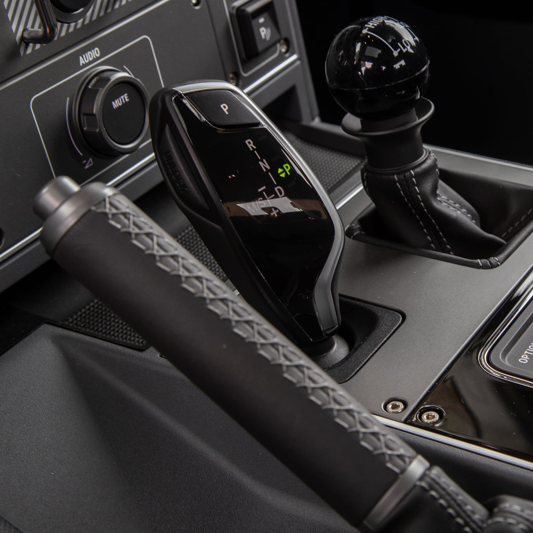 Close-up view of a car's gear shift area, featuring a modern gear selector, handbrake with stitched leather handle, and audio panel. Sleek and technical.