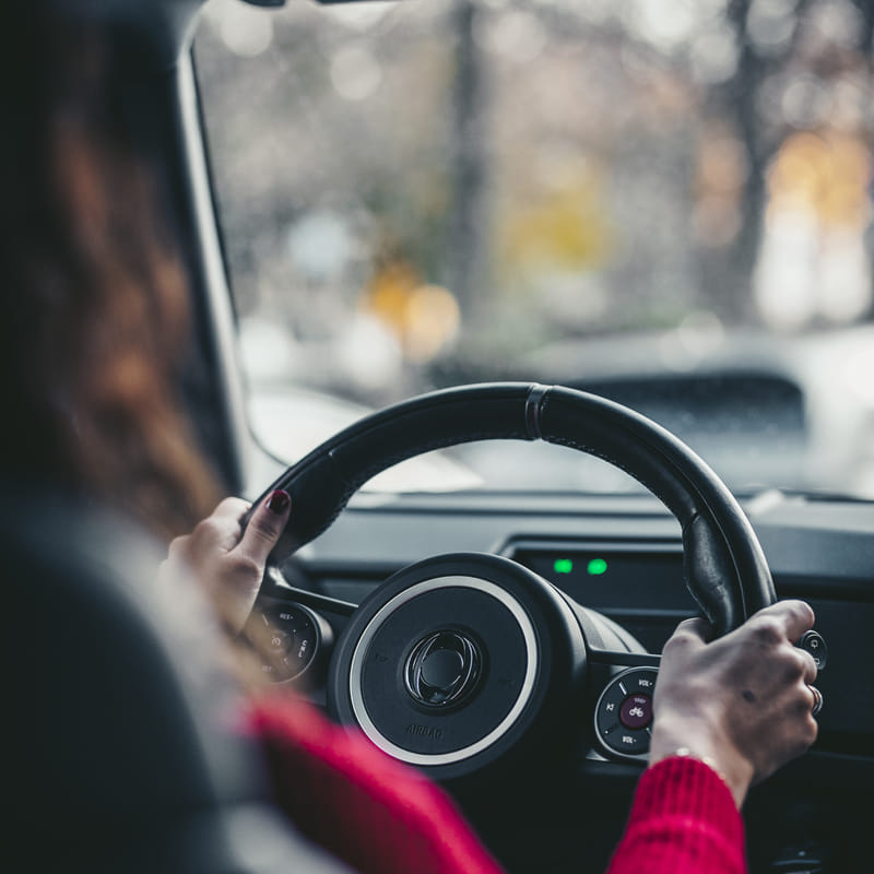 A close-up, driver-perspective shot of the Ineos Grenadier’s unique two-spoke steering wheel. A person with red fingernails and a red sweater has their hands on the wheel, with a blurred view of trees visible through the windshield.