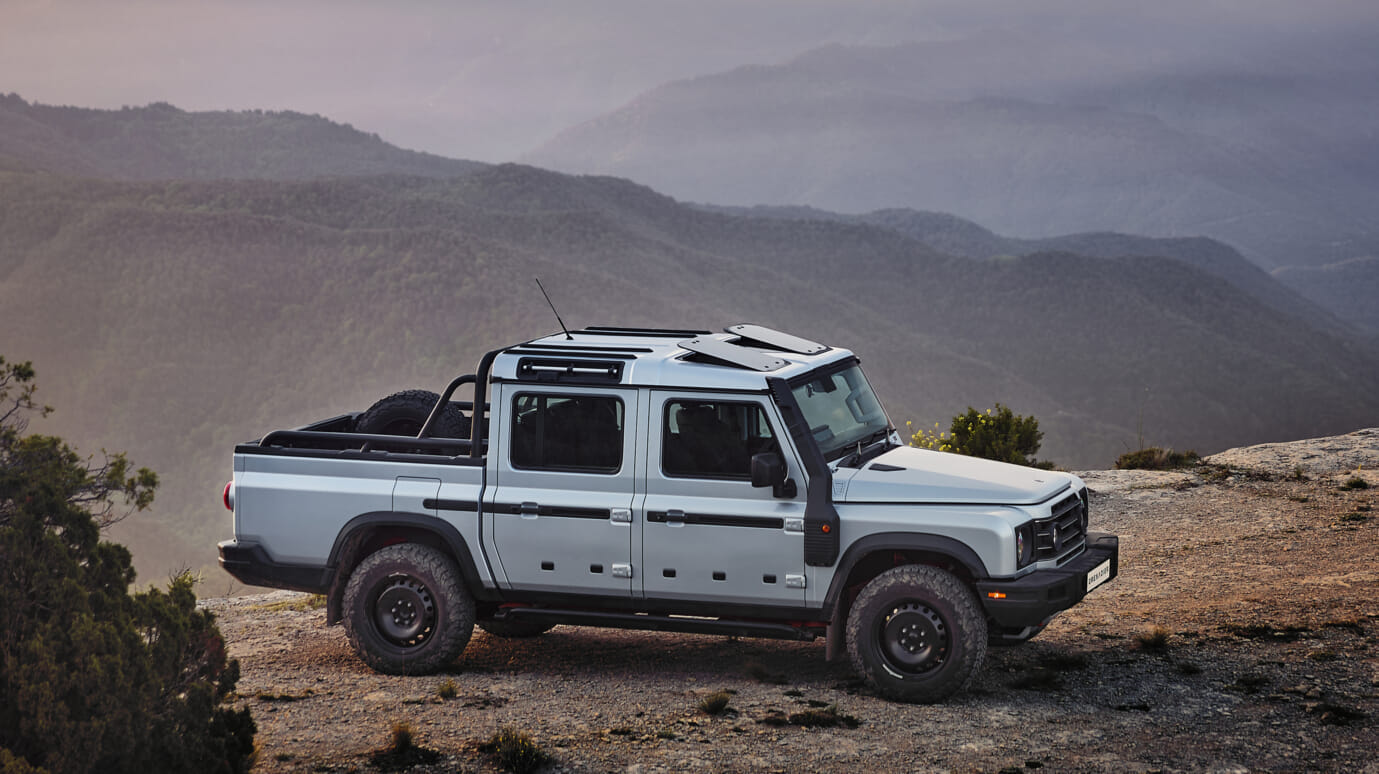 Ineos Grenadier Quartermaster is parked on a rocky hillside. The background features expansive, misty mountains at dusk.