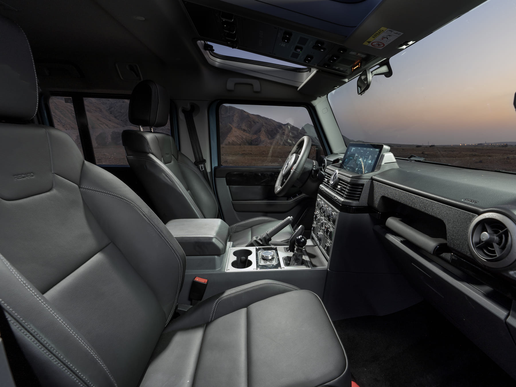 Modern SUV interior with leather seats and a sleek dashboard, featuring a digital display. The setting sun casts a warm glow over a mountainous landscape outside.
