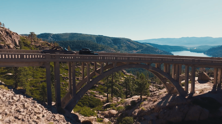 A side view of a dark INEOS Grenadier Station Wagon with a rooftop cargo box driving across a historic concrete arch bridge, with rugged mountains and a lake in the background.
