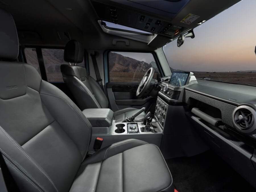 Modern SUV interior with leather seats and a sleek dashboard, featuring a digital display. The setting sun casts a warm glow over a mountainous landscape outside.