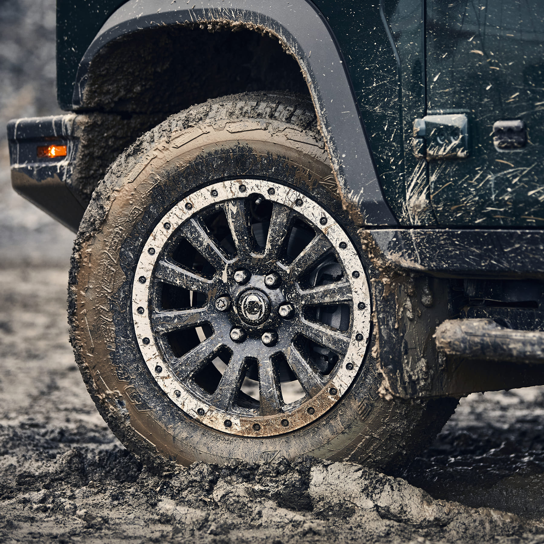 Close-up of a Jeep's front tire caked in mud, highlighting rugged off-road capability. The vehicle is dark green, conveying a sense of adventure.