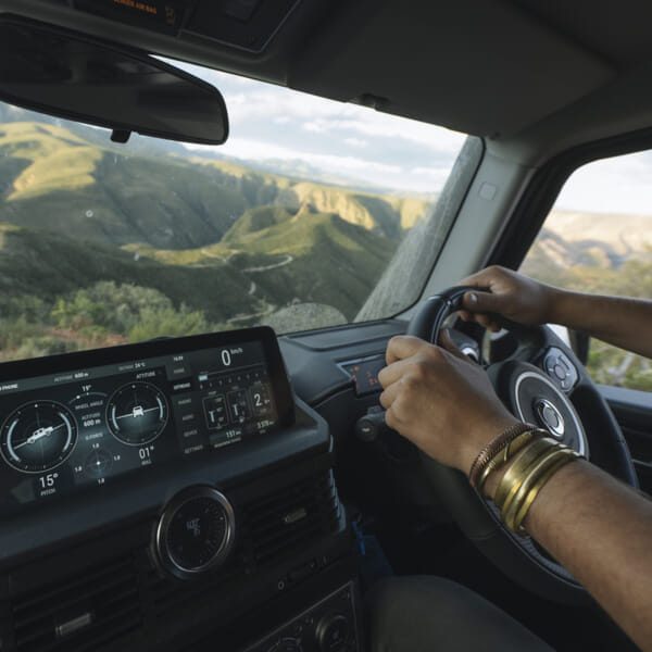 A person wearing gold bangles drives a vehicle through scenic mountain roads. The dashboard displays navigation info. The mood is adventurous and serene.