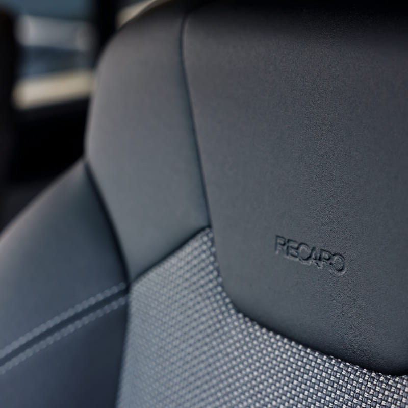 Close-up of a car seat headrest featuring the 'RECARO' logo, showcased with sleek black leather and textured fabric, conveying luxury and comfort.