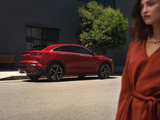 Red SUV parked on a sunlit street beside a tree, with a blurred woman in an orange dress walking by. The scene conveys a sense of elegance and urban style.