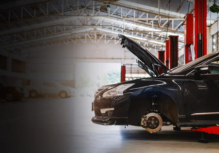 A black car is lifted in a spacious garage, with its hood open and front wheel removed, spotlighting maintenance work. Red lifts and cars line the background.