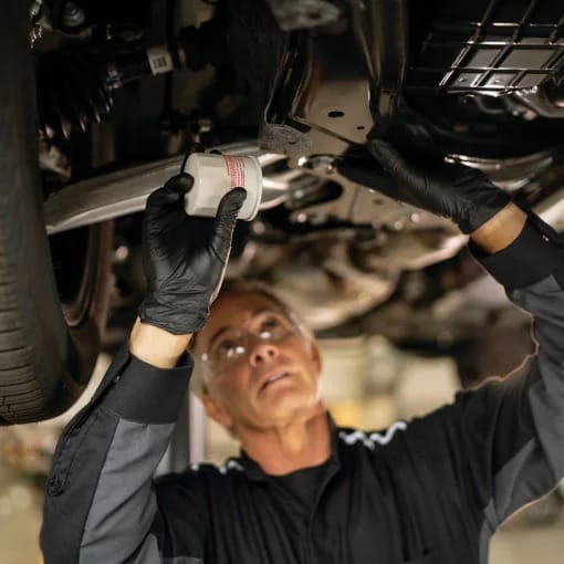 A mechanic wearing black gloves and a uniform inspects a car's underside, holding a tool with focus and precision in a well-lit garage.