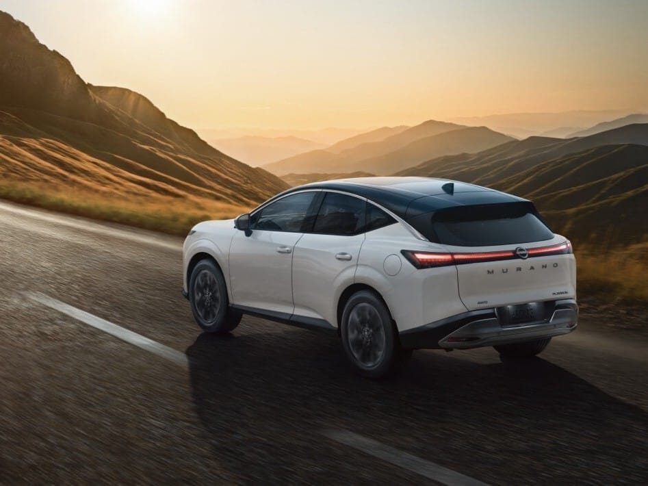 A white 2026 Nissan Murano driving on a winding road, with mountains and a sunset in the background, showcasing its sleek design.
