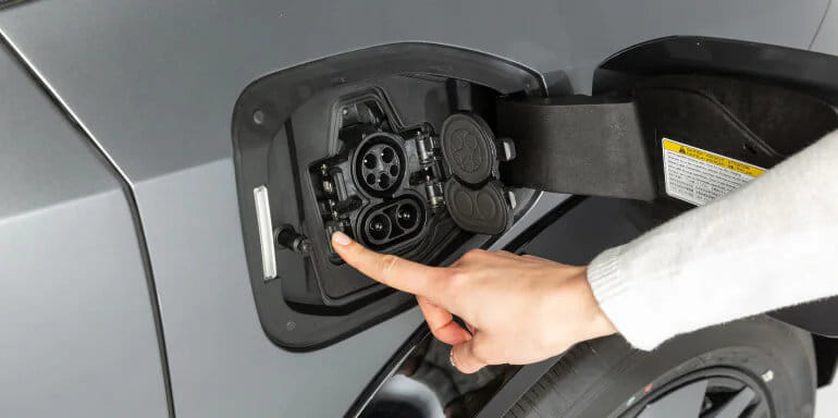 A person's hand points to an open electric car charging port on a gray vehicle, indicating interaction with eco-friendly technology.