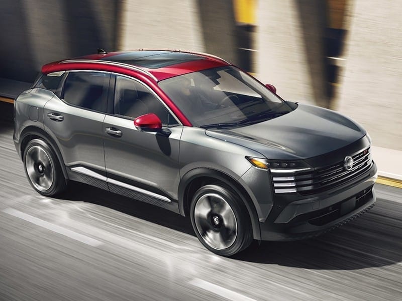 A sleek gray 2026 Nissan Kicks with a bold red roof and accents drives on a modern urban road, showcasing a dynamic design.