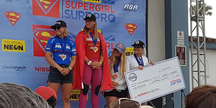 Four female surfers stand on a podium at the Supergirl Surf Pro event. One wears a red cape and holds a large check. They all wear medals, looking proud.