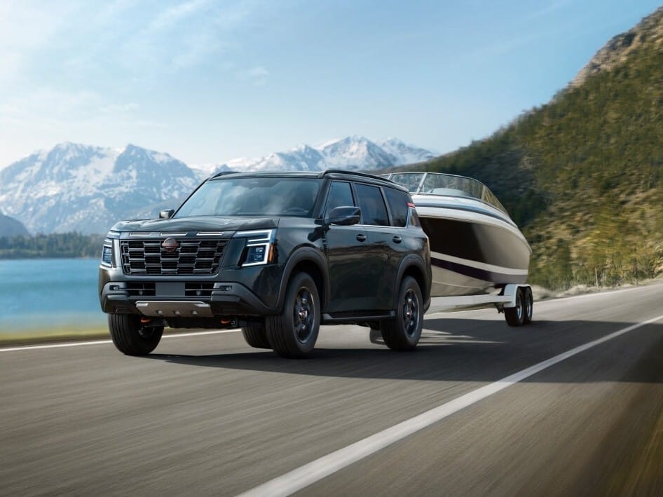 A black 2026 Nissan Armada tows a sleek boat along a scenic road with mountains and a lake in the background. Clear skies enhance the outdoor adventure vibe.
