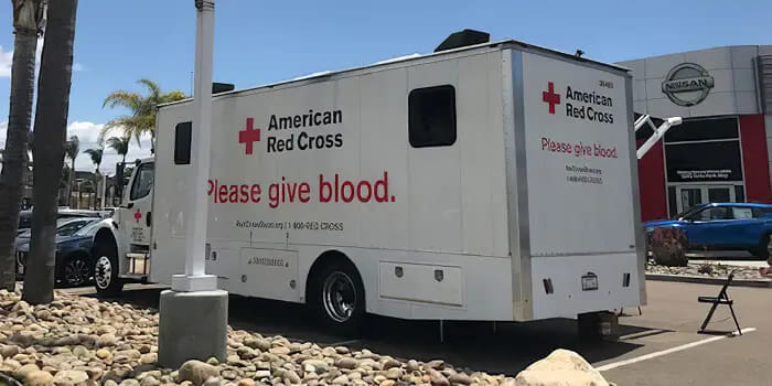 Mobile blood donation center with American Red Cross branding parked near a Nissan dealership. The text on the vehicle reads 'Please give blood.'