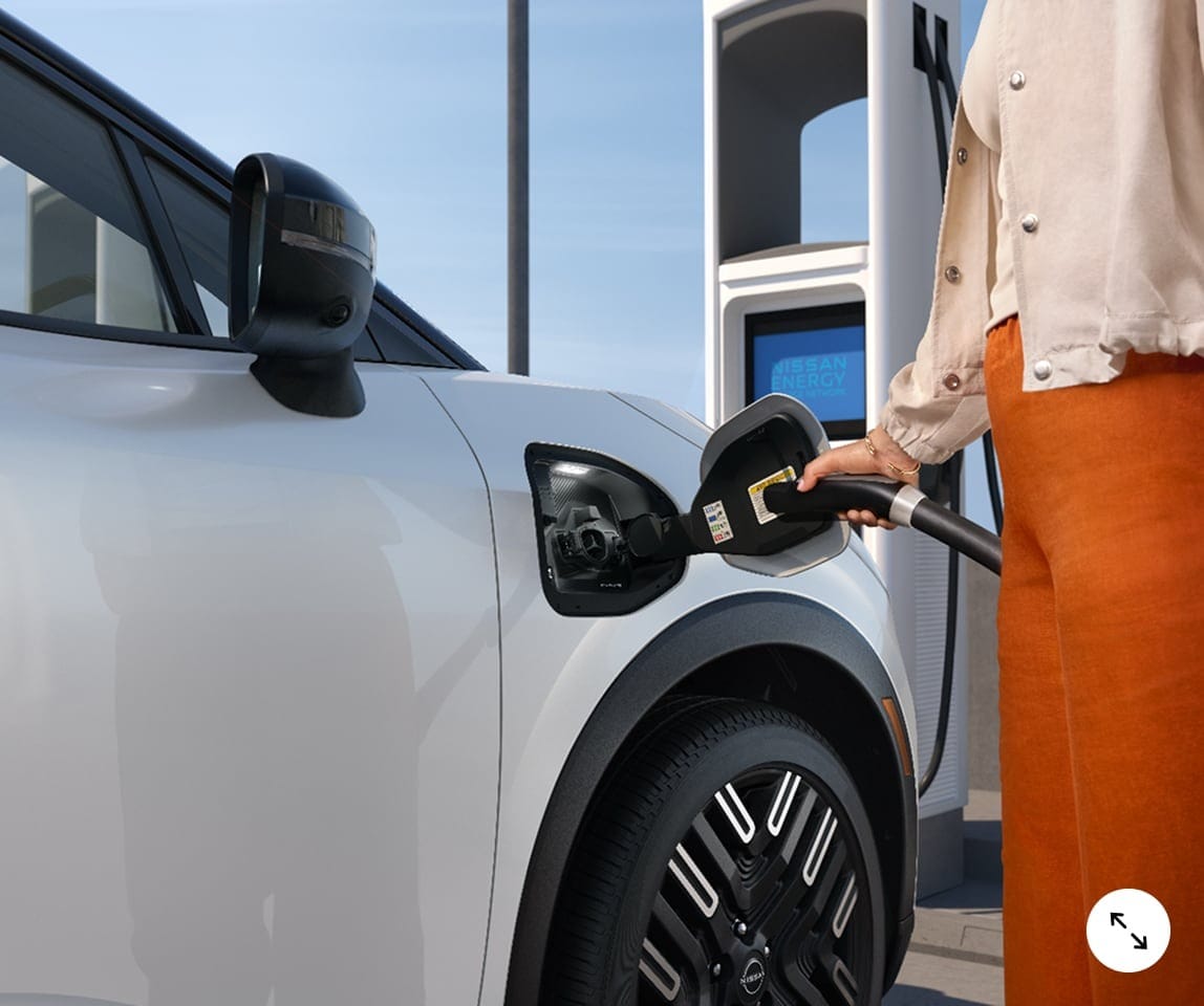 A person connects an electric charging cable to the 2026 Nissan Leaf at a charging station, with the station's screen visible in the background.