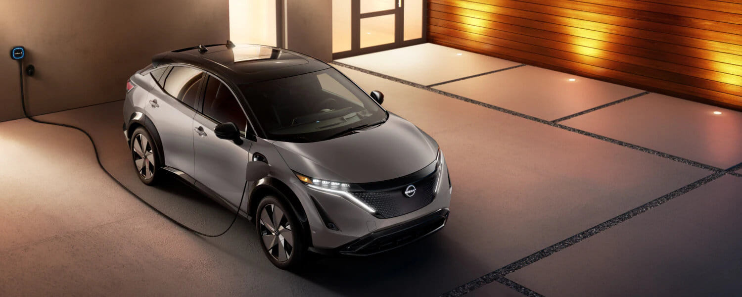 Sleek silver Nissan Ariya charging in modern garage with warm lighting. Car is plugged into wall, parked on smooth concrete floor, conveying futuristic vibe.