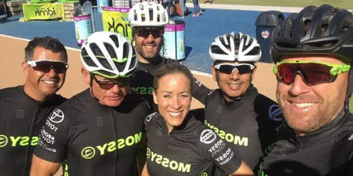 A group of six cyclists wearing matching black jerseys and helmets, smiling for a selfie outdoors on a sunny day. They appear happy and energetic.
