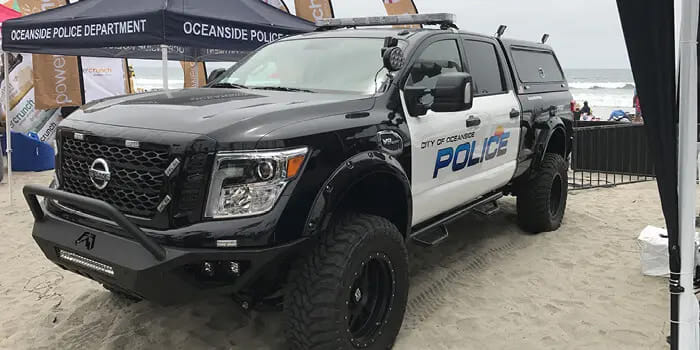A black and white police truck from the City of Oceanside on a sandy beach, parked under a tent labeled 'Oceanside Police Department,' with the ocean in the background.