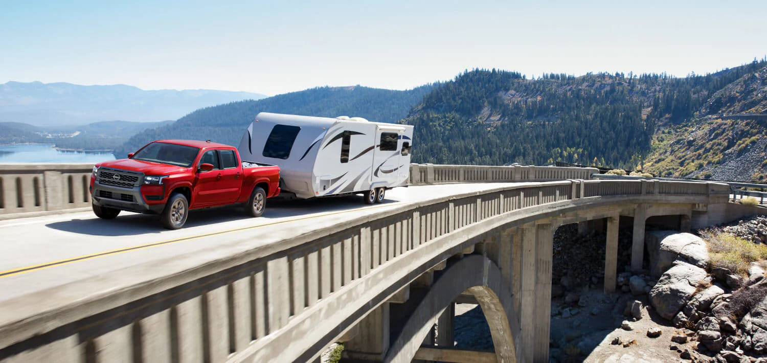 A 2026 Nissan Frontier PRO-4X towing a camper through a scenic forest, showcasing its powerful 310-hp V6 engine.
