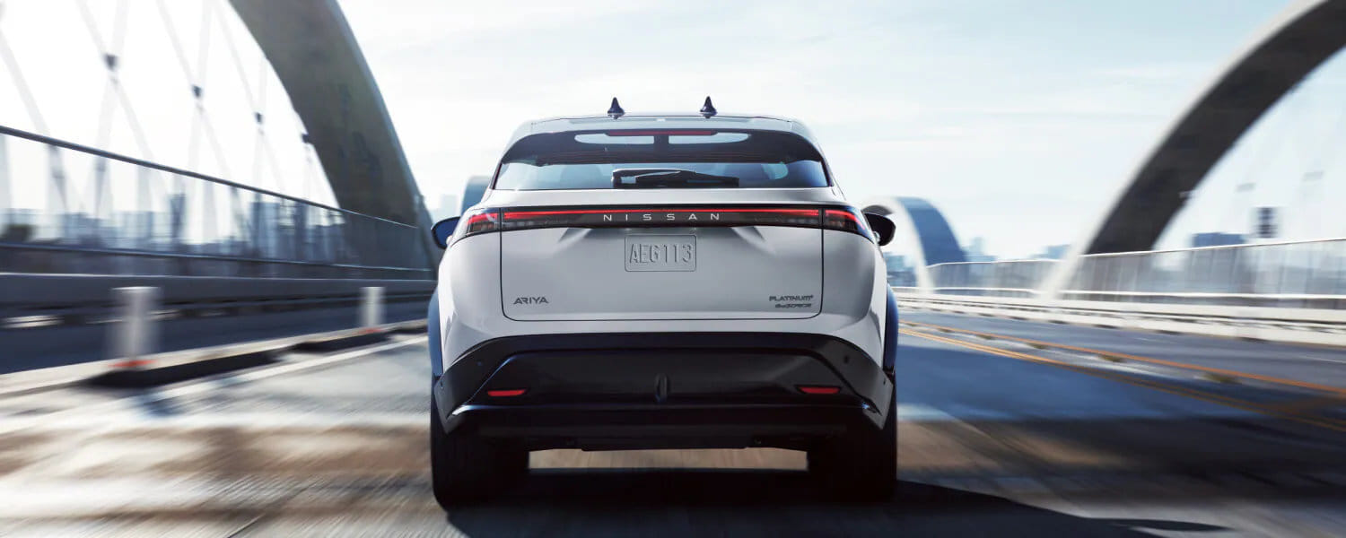Rear view of a white Nissan Ariya driving on a modern bridge. The sleek design suggests speed and innovation, with a bright, clear sky overhead.