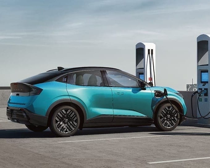A sleek turquoise 2026 Nissan Leaf is parked at a charging station, with the charging cable connected, against a clear blue sky.