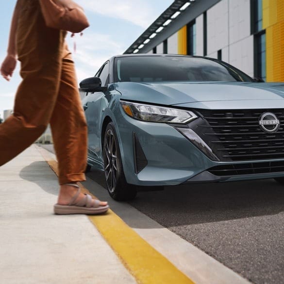 A person in brown pants and sandals walks past a parked blue Nissan sedan. The modern building in the background has a sleek design. The scene feels urban and dynamic.
