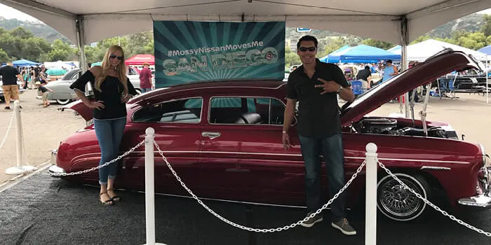 Man and woman posing with a vintage red car under a tent at a car show. The car's hood is open, sign reading '#MossyNissanMovesMe San Diego' behind.