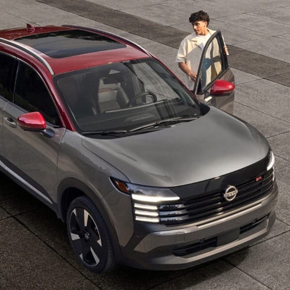 A man stands by an open door of a modern gray SUV with a red roof, parked on a stone surface. The car features sleek, contemporary styling.