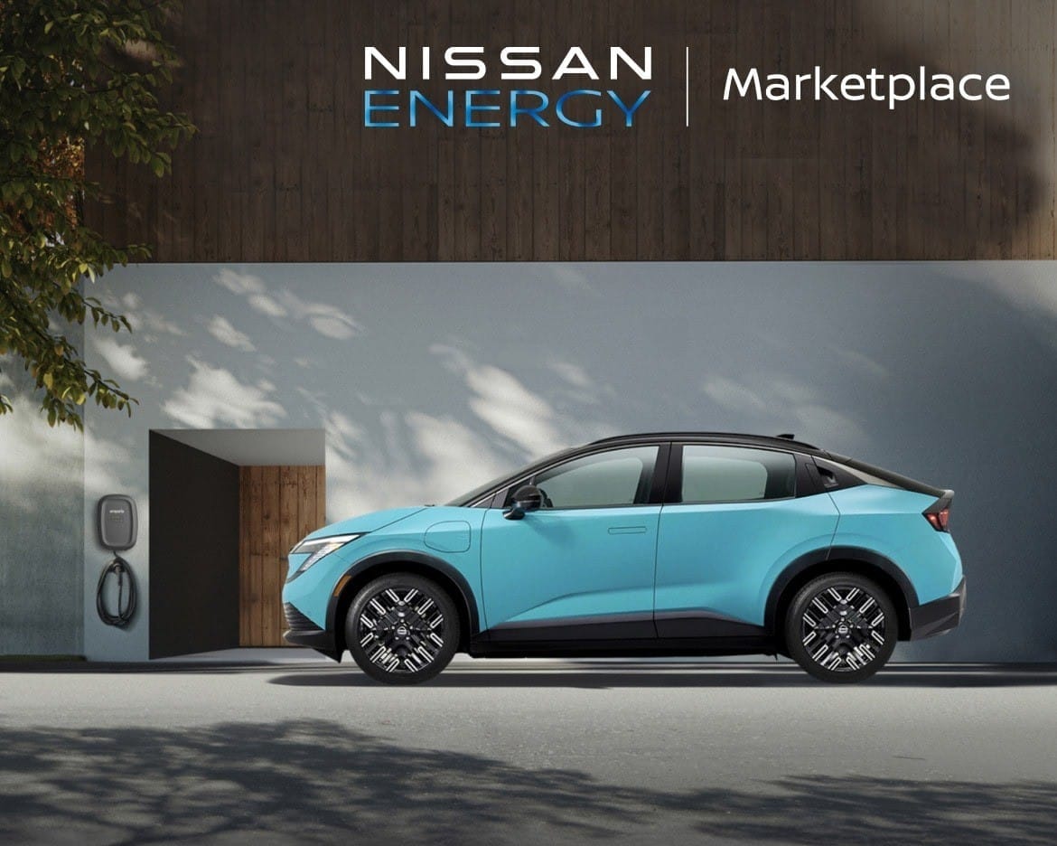 A blue electric car is parked beside a modern building with a charging station. Sunlight casts tree shadows on the wall, creating a serene ambiance.