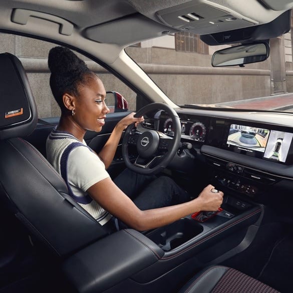A woman smiles while using the controls inside a modern car. The dashboard displays advanced tech features. The scene conveys comfort and enjoyment.