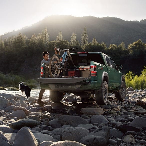 A person unloads a bicycle from a green pickup truck in a rocky riverside setting at sunrise. A dog stands nearby. The scene suggests adventure and tranquility.